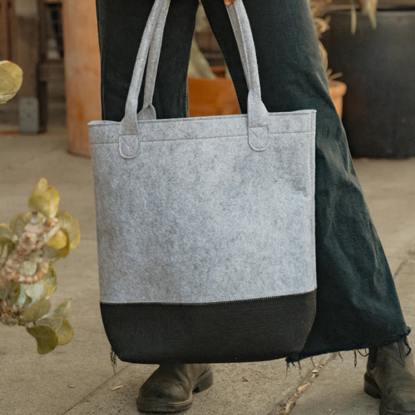 Person holding a stylish gray and black felt tote bag with long handles over dark pants indoors.