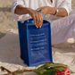Hand holding a blue eco-friendly tote made from 100% ocean plastic on a beach blanket with flowers nearby