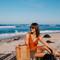 Woman sitting on a beach with the Walrus Cooler, wearing sunglasses and an orange top.