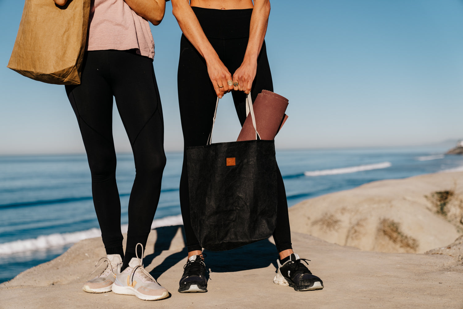 Two people wearing black leggings and sneakers standing on a rocky shore with the ocean in the background