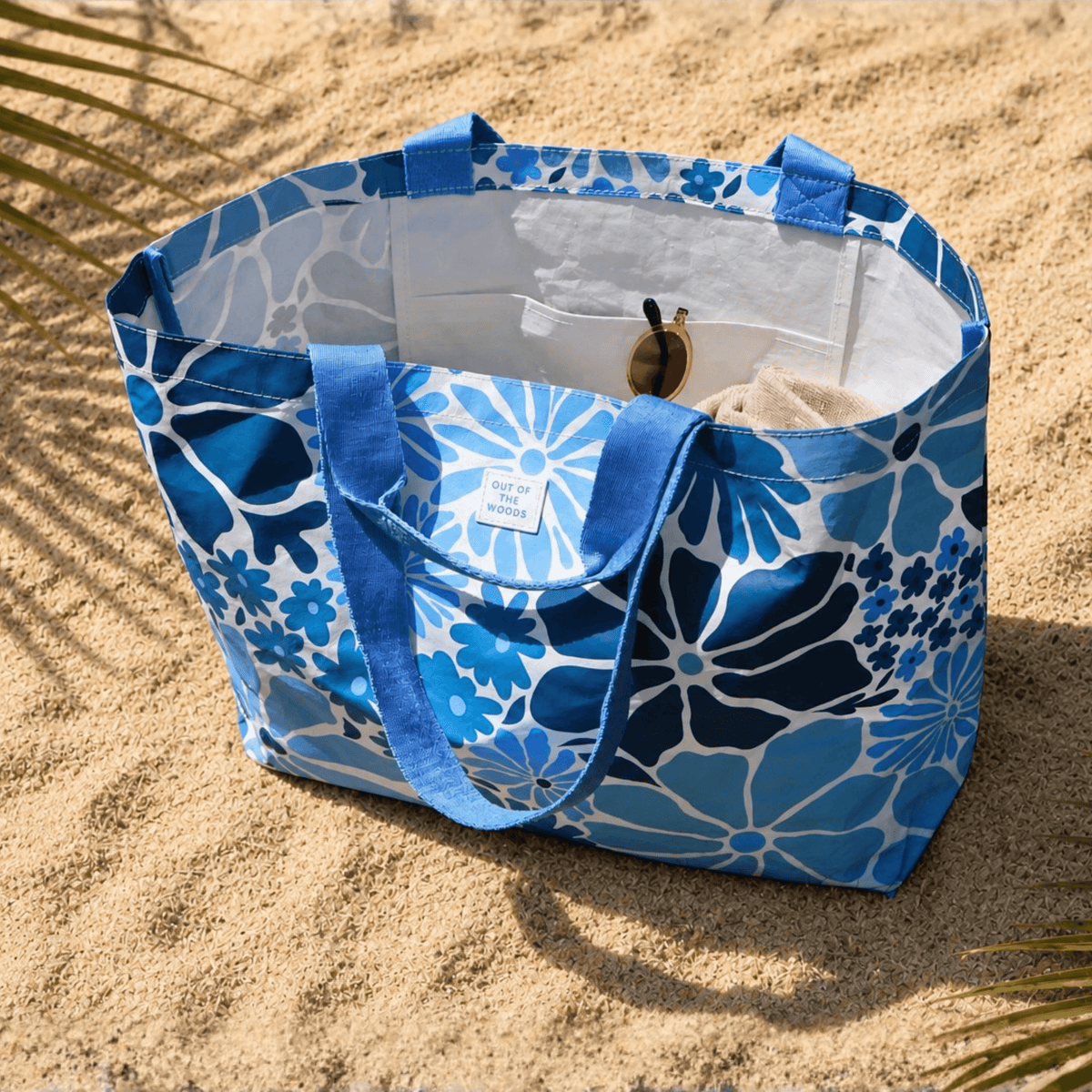 A blue and white floral tote bag with blue shoulder straps sits on sandy ground outdoors, with palm leaves casting shadows nearby. A small fabric brand tag is visible on the front, and a small accessory is tucked into the bag's interior pocket.