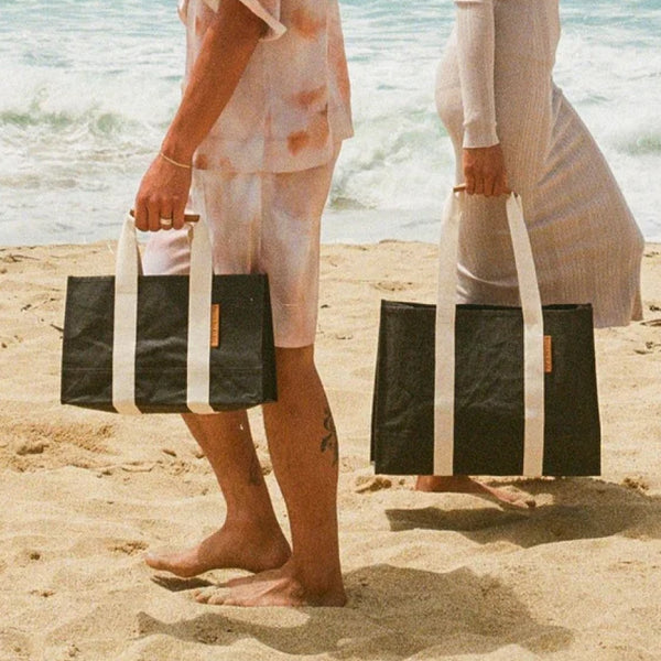 Two people holding black tote bags with white straps walking barefoot on a sandy beach near the ocean