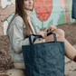 Woman holding a blue washable paper Iconic Shopper tote bag in front of a colorful mural.