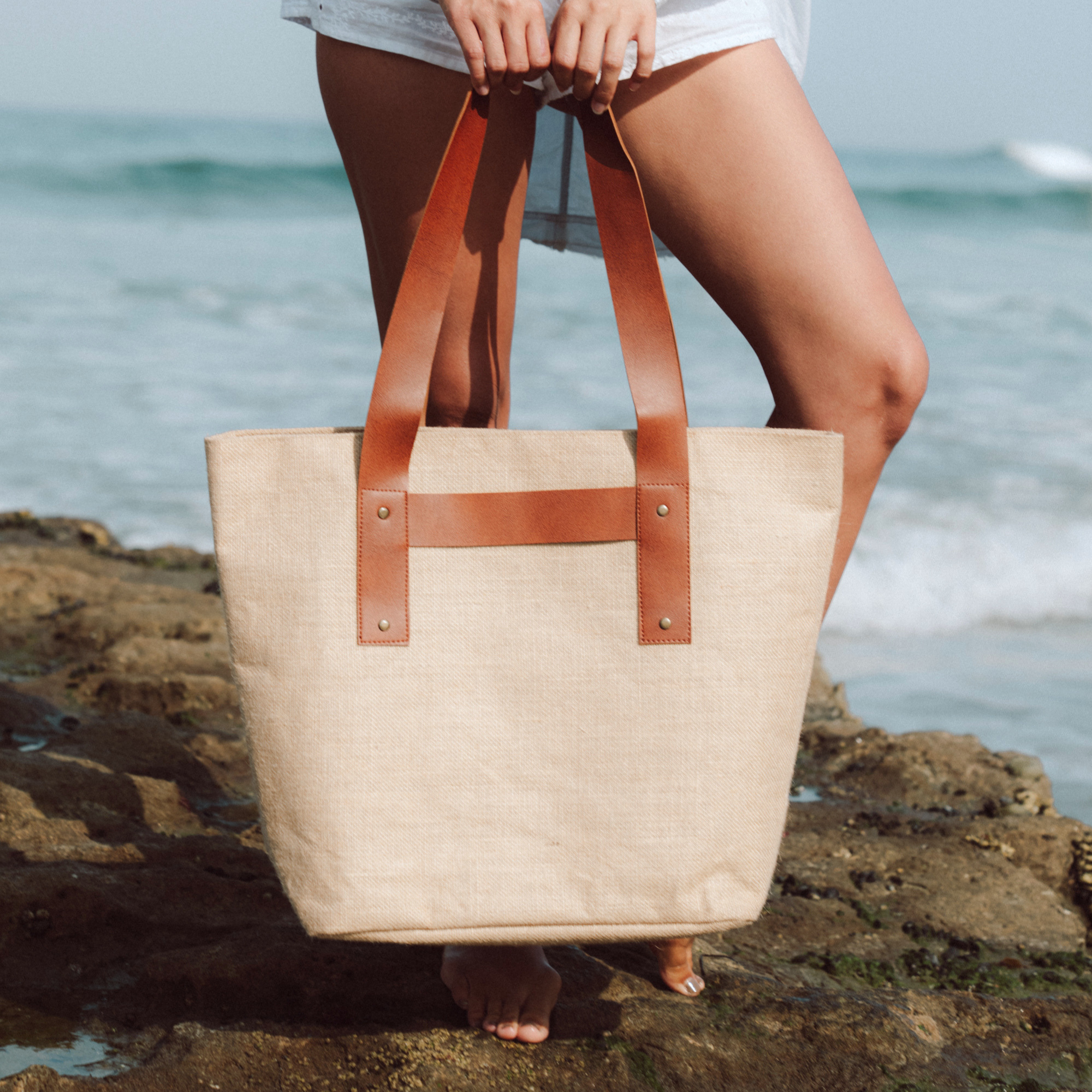 A jute tote bag with brown vegan leather handles and trim, held by a person standing on rocks with the ocean in the background.