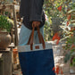 Person carrying blue and natural tone Jute Tall Tote with leather handles in outdoor garden market setting