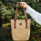 Person holding a beige tote bag with brown straps in a forest setting