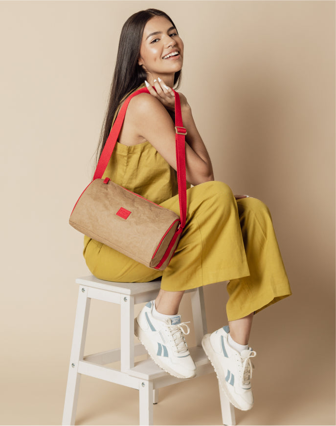 Woman wearing yellow outfit sitting on white stool holding tan and red shoulder bag with white sneakers