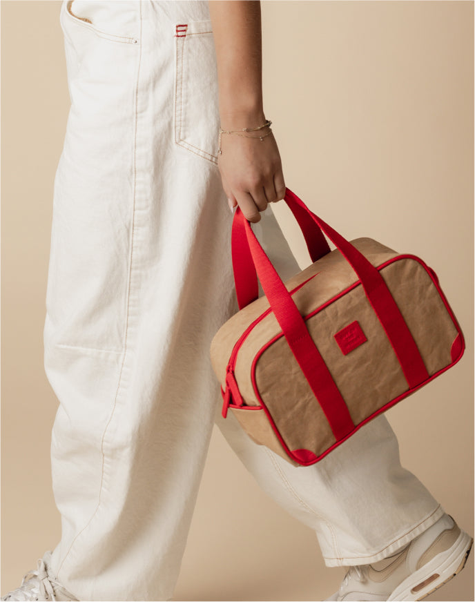 Person holding a small beige and red duffel bag against a neutral background with white pants and sneakers