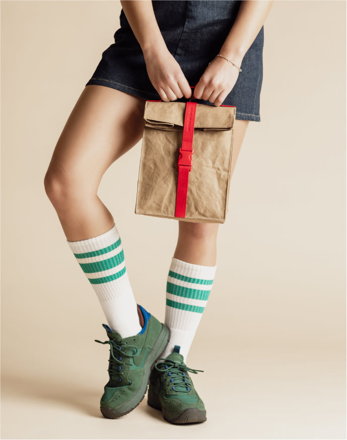 Person wearing green sneakers and striped socks holding a beige and red fabric bag against a neutral background
