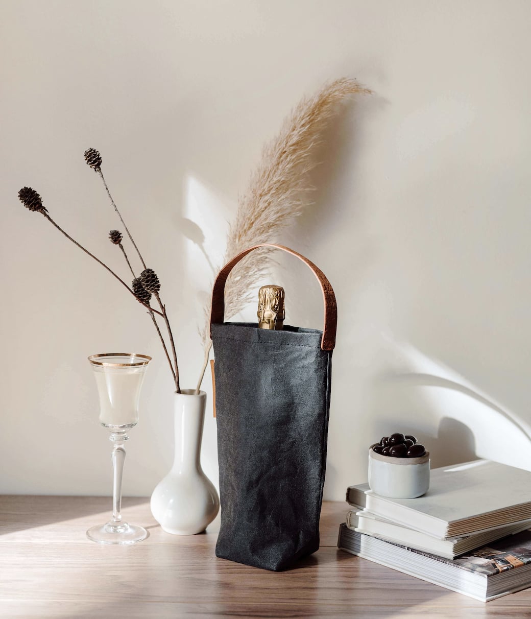 Bottle wrapped in a black fabric holder with a leather handle on a table with vases and stacked books nearby