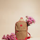 Zip Around Sling in beige with red accents, compact and water-resistant, shown with pink flowers against neutral background